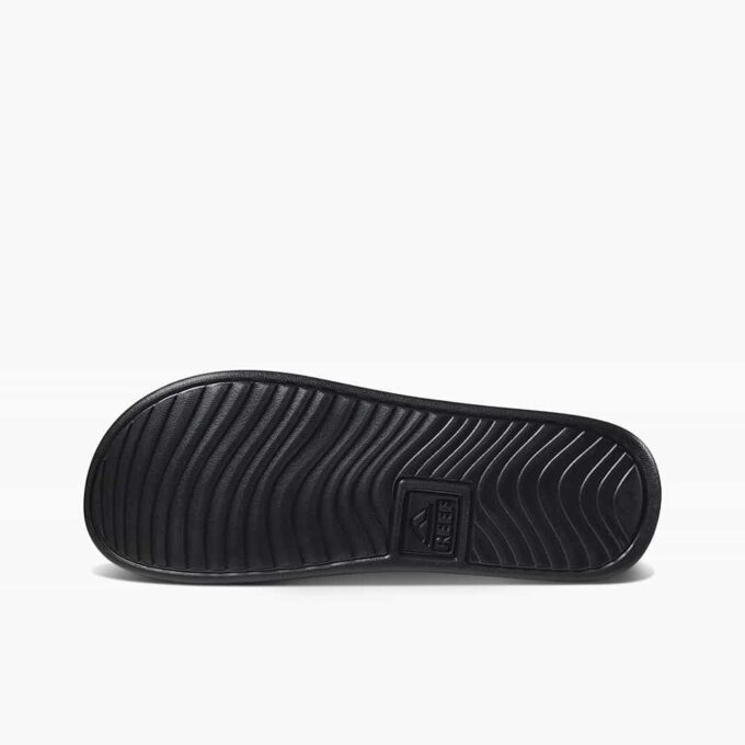 "Bottom view of the black sole of a Reef CI4060 slide sandal showing wavy tread pattern and embossed Reef logo in the center."