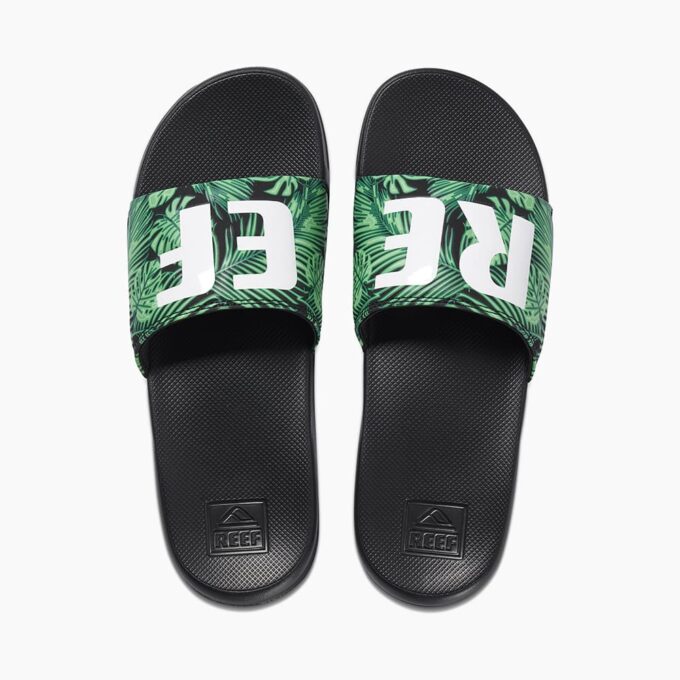 "Top view of a pair of Reef CI4060 slide sandals with black footbeds and green tropical leaf-patterned straps featuring the white 'REEF' logo."