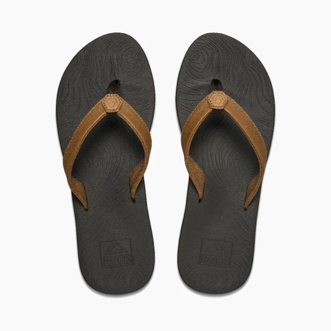 "Pair of black flip-flops with brown suede straps, viewed from above, featuring a textured footbed with wave-like patterns and the Reef logo embossed near the heel."