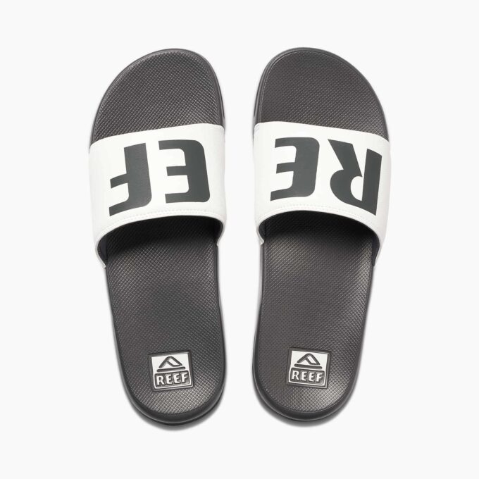 "Top view of a pair of Reef RF0A3ONDGRW slide sandals featuring black footbeds with textured surface and white straps displaying bold black 'REEF' branding across both sandals."