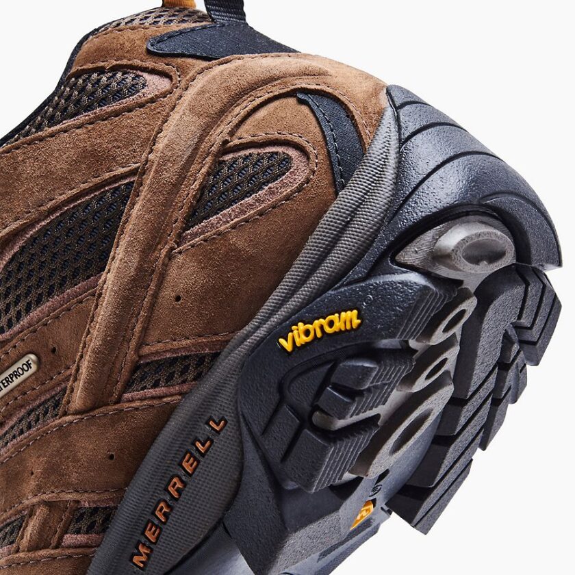 "Close-up view of the heel and sole of a brown Merrell J06051 hiking boot, showcasing suede and mesh materials with waterproof labeling, black and grey Vibram outsole, and branded "Merrell" and "Vibram" logos in orange and yellow respectively."