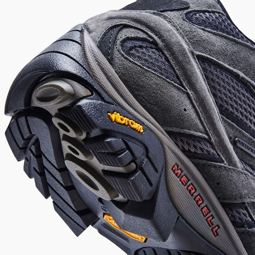 "Close-up image of the sole and side of a Merrell J06053 hiking shoe in dark gray suede and mesh. The shoe features a Vibram outsole with black and gray tread and the "Merrell" brand name in red on the side midsole. A small tag on the side indicates the shoe is waterproof."