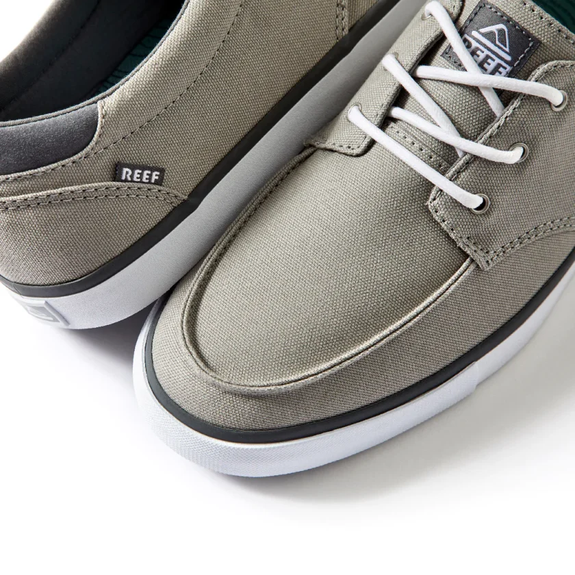 "Angled close-up of Reef RF0A3FE4GRW casual sneakers in gray, featuring white laces, moc toe stitching, canvas upper, and black-and-white rubber outsole with visible Reef branding tag."