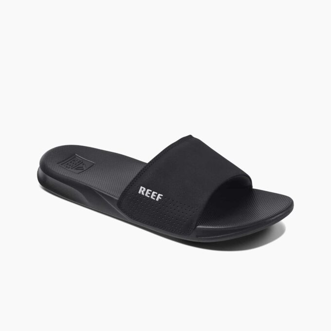 "Angled side view of a black Reef RF0A3ONDBLA slide sandal with a perforated strap featuring the white Reef logo and a textured footbed."