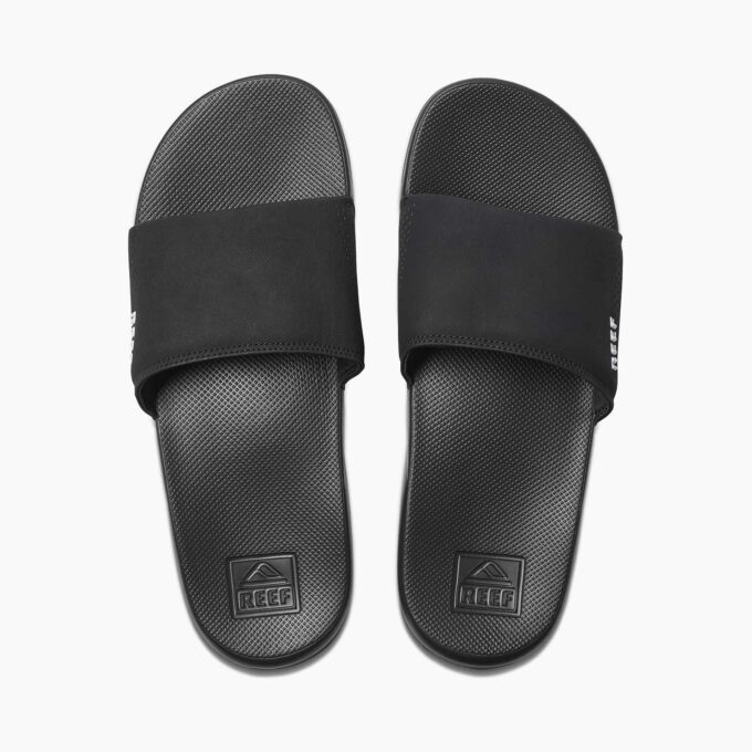 "Top view of a pair of black Reef RF0A3ONDBLA slide sandals showing textured footbeds, perforated straps, and embossed Reef logos near the heels."