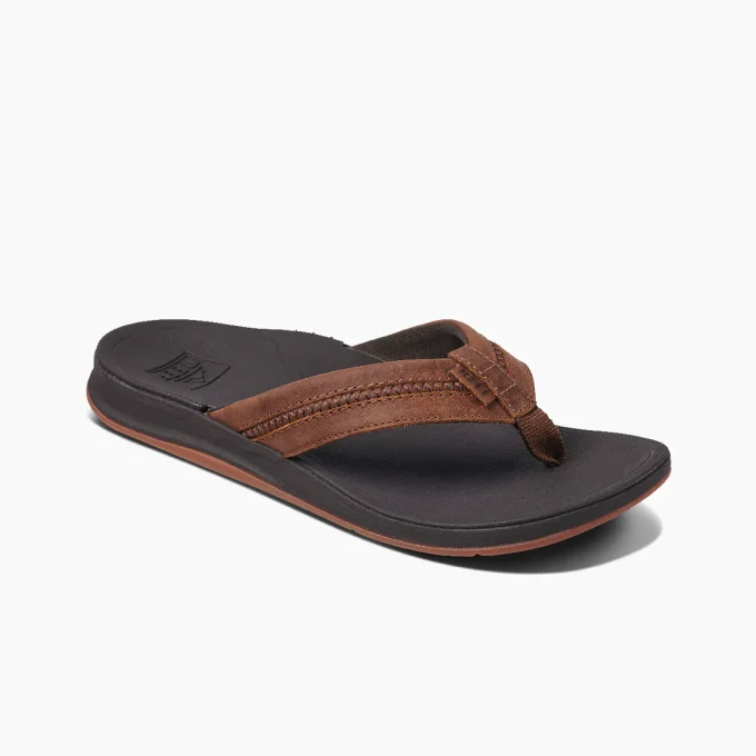 "Angled view of a brown leather Reef flip-flop with a cushioned dark footbed and brown rubber outsole, showing detailed stitching on the straps."