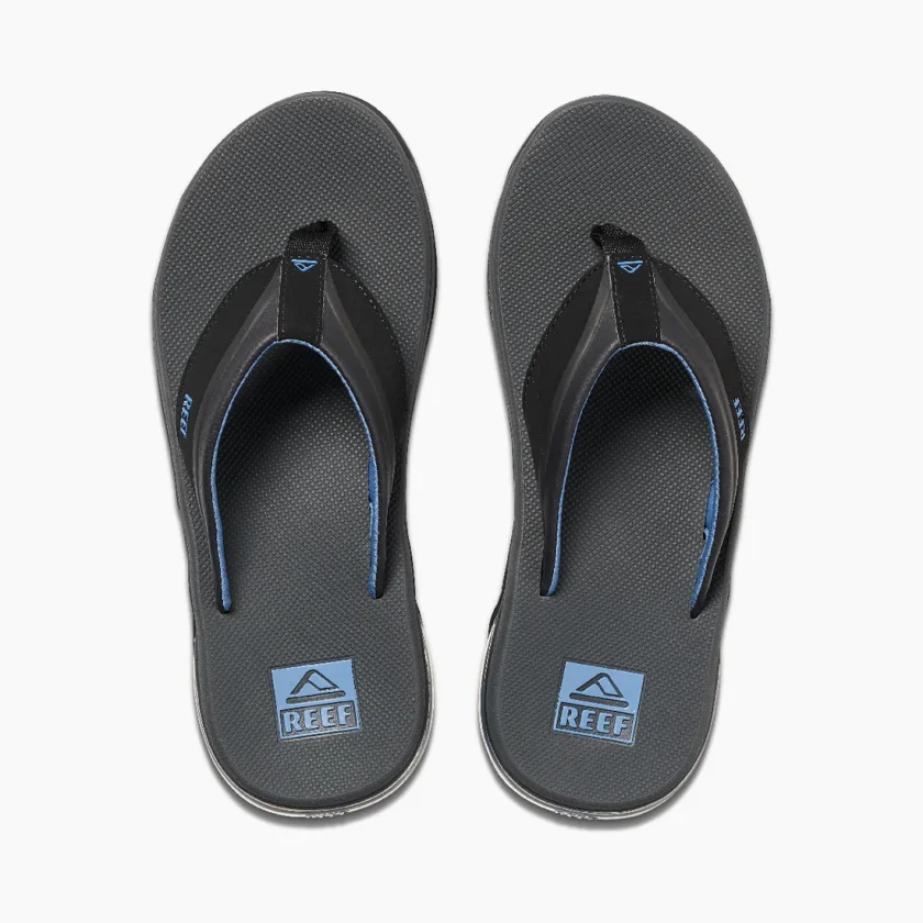 "Top view of a pair of Reef RF0A3YMIGBL sandals featuring black straps with blue lining, textured dark gray footbeds, and blue Reef logos printed near the heel area."