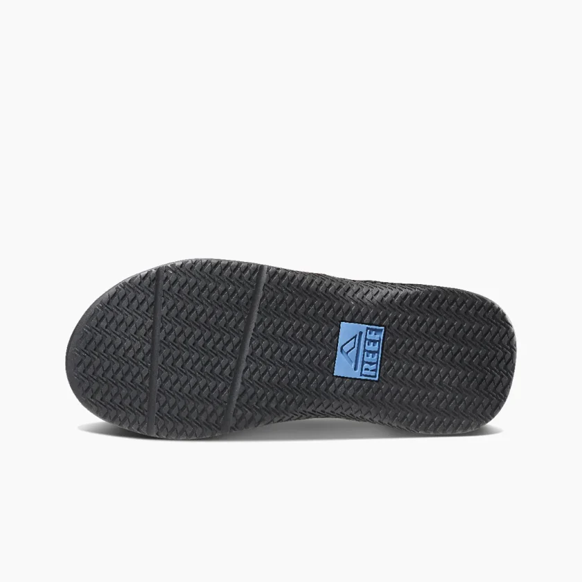 "Bottom view of the Reef RF0A3YMIGBL sandal displaying a black rubber outsole with a herringbone tread pattern and a blue Reef logo positioned at the midfoot for traction and branding."