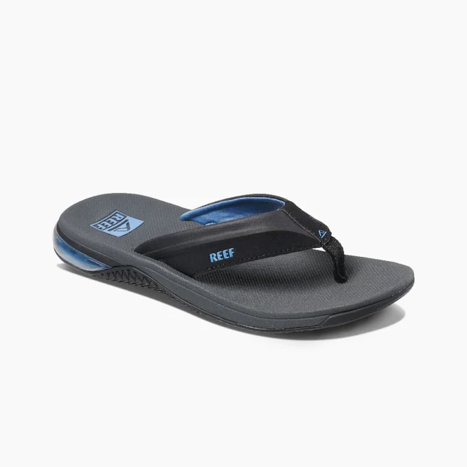 "Angled view of a Reef RF0A3YMIGBL sandals featuring black straps with blue lining, textured dark gray footbeds, and blue Reef logos printed near the heel area."