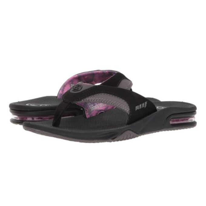 "Angled front view of Reef Fanning sandals (model RF001626BLG) in black and grey with purple patterned lining. Features include a cushioned footbed, soft fabric thong strap with contrast stitching, visible air cushioning in the heel, and the signature built-in bottle opener on the outsole."