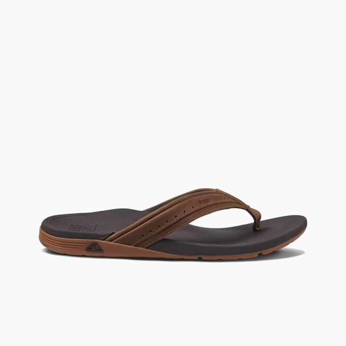 "Side profile of a brown and black Reef flip-flop sandal with a cushioned footbed and leather-like straps." "Single brown thong-style sandal from Reef, featuring a contoured black footbed and brown sole with embossed logo." "Brown Reef flip-flop sandal with perforated straps and textured black footbed, isolated on white background."